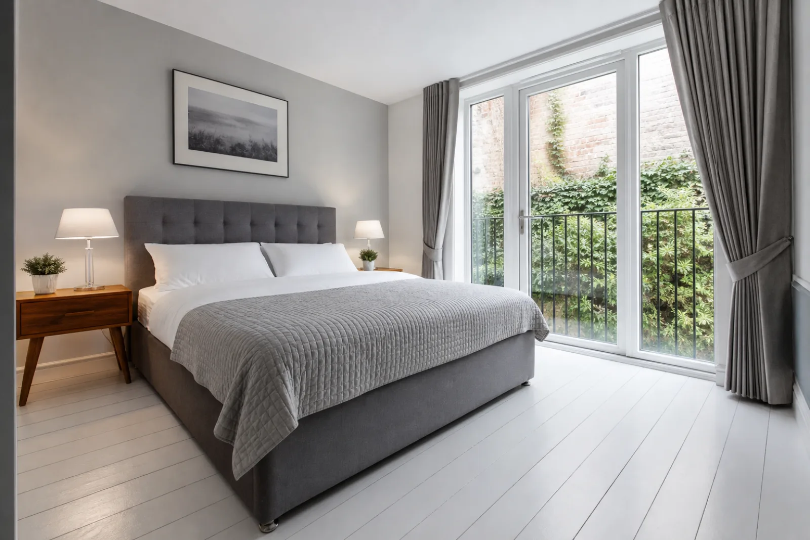 A freshly cleaned, spotless bedroom with painted floorboards and a neatly made bed in a British modern apartment with floor-to-ceiling windows and a Juliet balc
