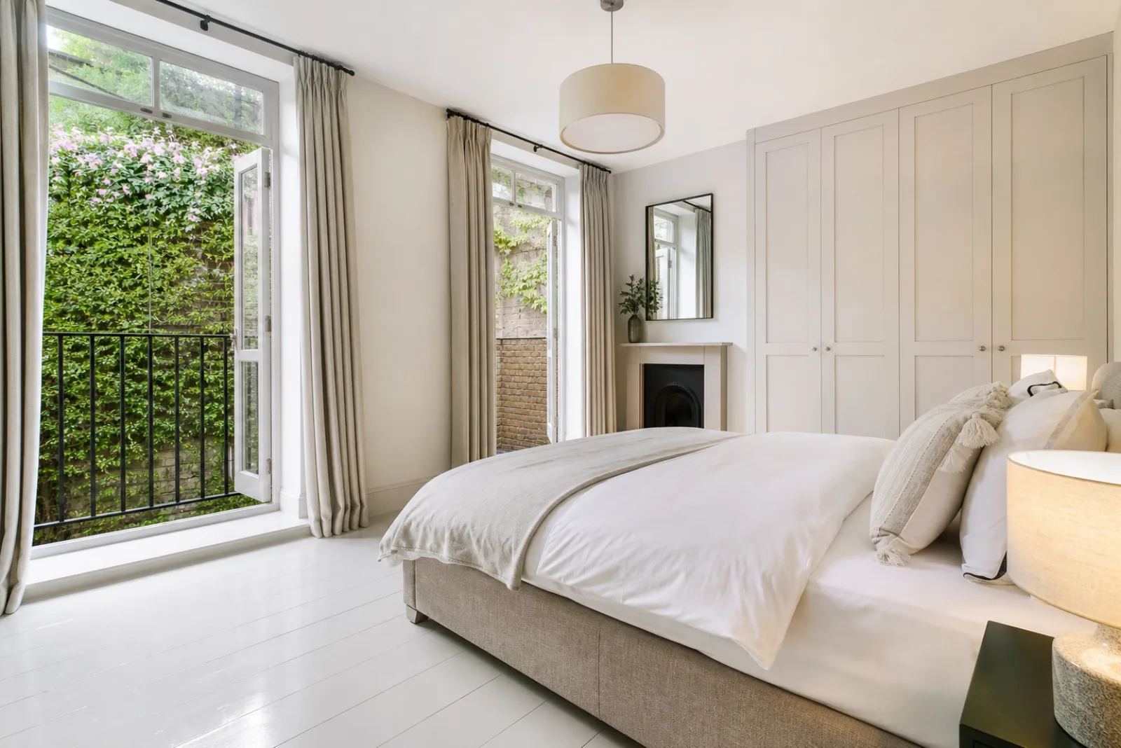 A freshly cleaned, spotless bedroom with painted floorboards and a neatly made bed in a British modern apartment with floor-to-ceiling windows and a Juliet balc
